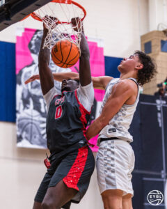 Two basketball players in youth travel basketball compete as one dunks in the hoop.